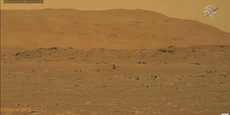 Au centre de cette première image en très basse définition transmise tout à l'heure depuis Mars à 278 km de distance, on distingue le minuscule hélicoptère Ingenuity, en vol, à quelque distance du sol. Crédit: Nasa / JPL Caltech. (Image agrandissable plein écran à l'intérieur de l'article)